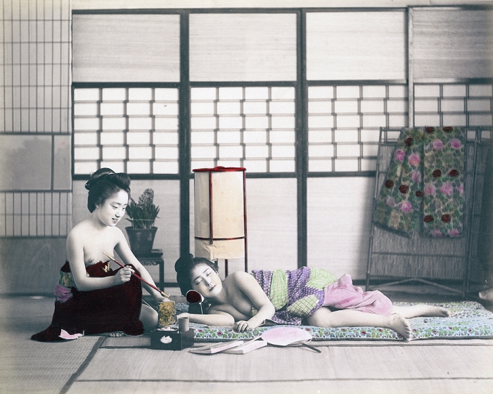 Woman sleeping on a futon  1890s  A woman is lying half naked on a  futon , her head is resting on a high pillow  hakomakura  to protect her hairstyle. There is a tobacco tray, two books and a fan in front of her and an  andon  lamp behind her. The woman next to her is smoking a long  kiseru  pipe. On the far right is a screen or clothes rack with what appears to be an  obi , used to tighten a kimono. Albumen photograph, 1890s.