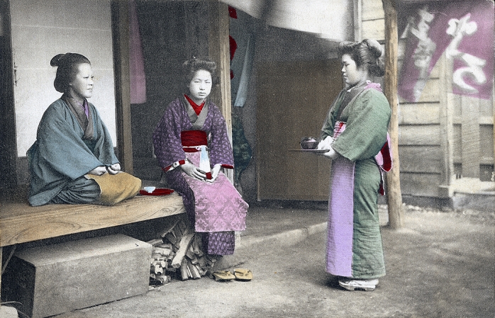 Teahouse  Date of photograph unknown  A maid servant in kimono, apron and traditional hairstyle at a tea house. She is holding a tray with a bowl. Two women sit on the engawa  porch .