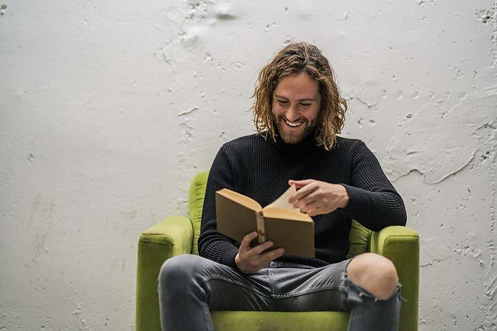 masculine gender Smiling young man reading book while sitting on armchair against wall at home