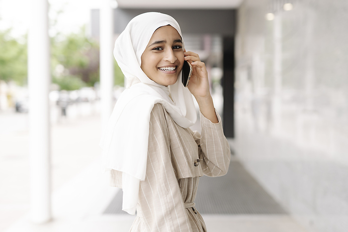 female Muslim teenage girl smiling while talking through smart phone in city