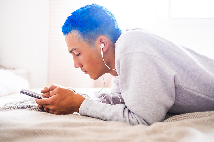 Young man with blue hair using smartphone and listening to music lying on bed