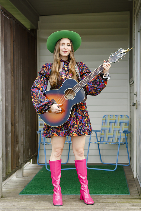 A stylish woman with long hair and acoustic guitar stands on porch