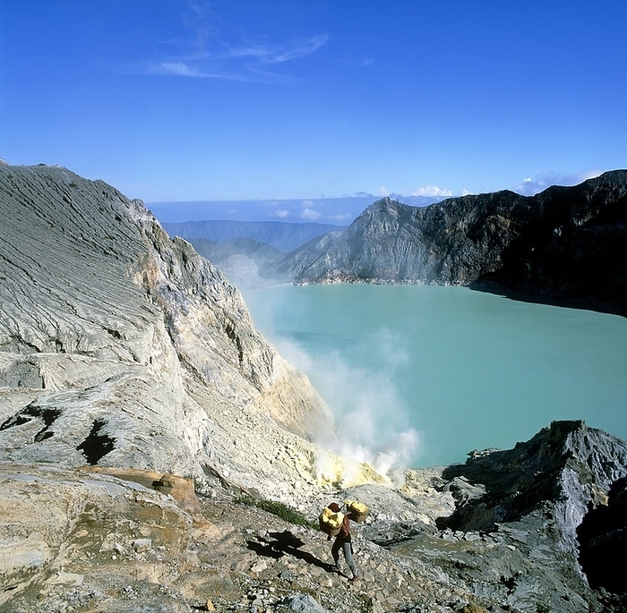 Volcanic crater lake with sulphur deposits Volcanic sulphur mining. View of a man carrying two baskets of sulphur after taking it from deposits around a crater lake at Kawah Ijen volcano, Java, Indonesia. Elemental sulphur is produced around volcanoes as hot sulphur  containing gases from volcanic vents condense and form deposits when they contact the cooler air. A sulphurous vent can be seen steaming behind the man at lower centre. Crater lakes such as this one are formed when a giant volcanic eruption leaves a crater which becomes filled with water. The water is typically hot, highly acidic and rich in sediment, which leads to its vivid colouration.