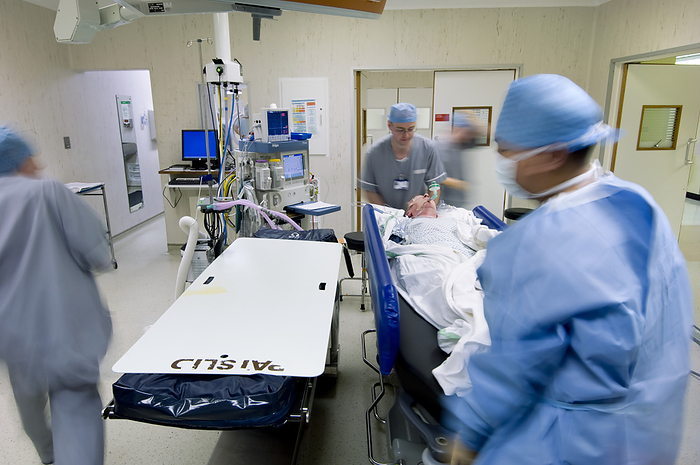Preparing for surgery MODEL RELEASED. Preparing for surgery. Surgical team preparing to lift a patient from a trolley bed onto an operating table. The patient has been anaesthetised, and the surgical team are wearing sterile clothing to help prevent infection of the patient. Surgical monitors and anaesthetic equipment are in the background at upper left. Photographed in the UK.