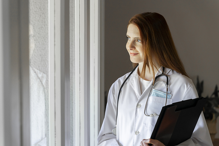female Thoughtful young female doctor smiling while looking through window in home office, Photo by VITTA GALLERY