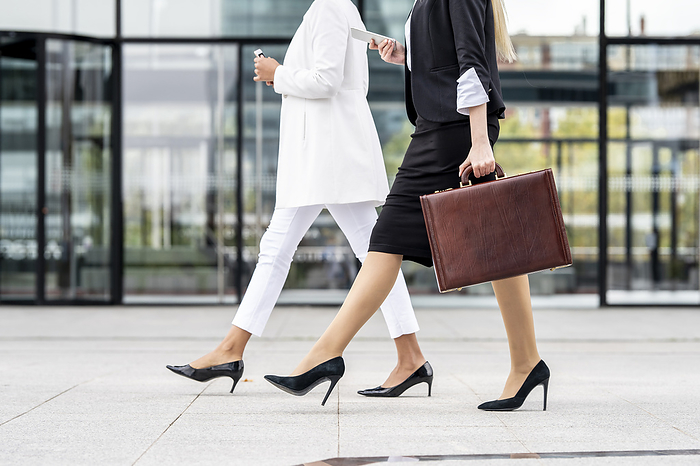 business Businesswoman with briefcase using mobile phone while walking by colleague on footpath, Photo by GER