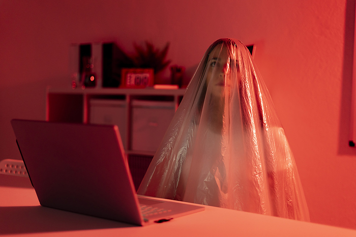 female Thoughtful woman covered in plastic sitting with laptop at desk in home office during pandemic, Photo by Giorgio Fochesato