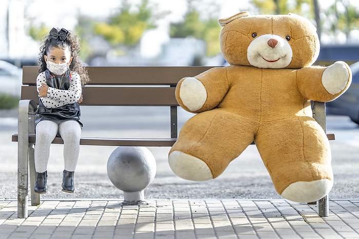 Girl wearing face mask sitting with teddy bear at social distance on bench