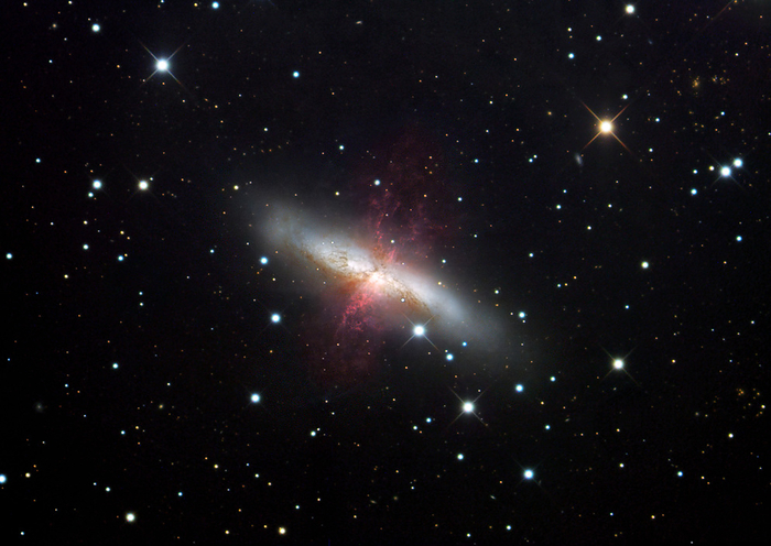 Cigar galaxy  M82  Cigar galaxy  M82 , optical image. This galaxy lies around 12 million light years away in the constellation Ursa Major. Its irregular shape is due to the distorting gravitational effects of its close neighbour, the larger galaxy M81  not seen . The interactions have stimulated star birth and death within M82 to around 10 times the normal level. The red regions are areas where radiation from hot young stars have ionised the surrounding hydrogen gas, causing it to glow. There is a strong stellar wind from the centre of the galaxy, caused by supernovae, or explosive star deaths.