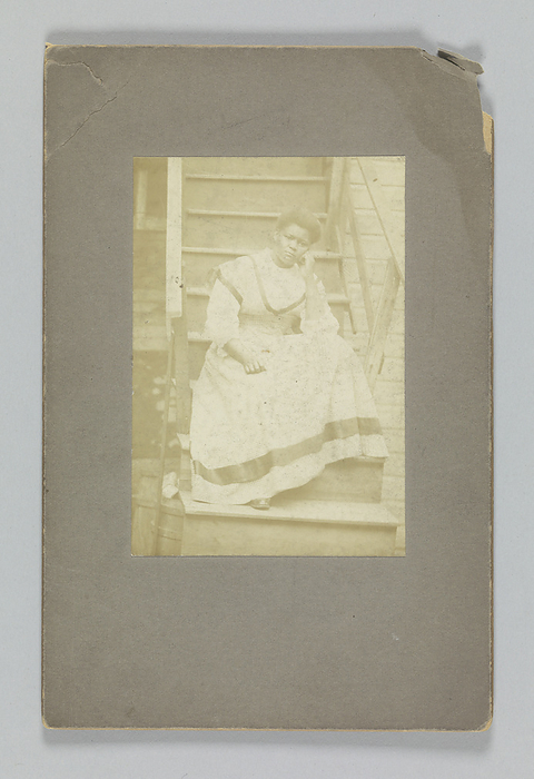 Albumen print of an unidentified woman sitting on stairs, 1850 1895. Creator: Unknown. A deep purple reddish brown photograph of an unidentified African American woman sitting on a set of wooden stairs. She is posed with her left arm resting on her left knee. Her head is resting on her left hand. Her right hand is placed on her lap. She is wearing a light colored dress with a dark stripe around the hem of the skirt. The stairs are attached to a building. The photograph is adhered to a dark gray mat. The back of the mat is blank