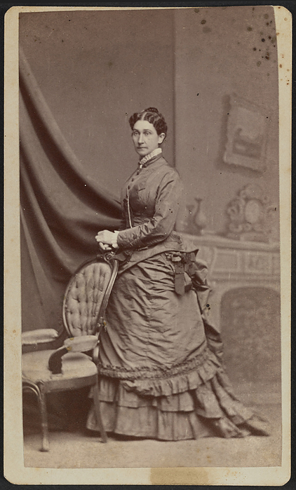 Carte de visite portrait of Anna M. Stanton, 1869 1877. Creator: Horning s Photographic Rooms. Carte de visite of Anna M. Stanton shown in full portrait. Stanton is standing with her body turned to the side and her left side foremost. Her head is turned to the camera. She wears her hair parted at the center and gathered into an updo behind her head. She has on a dark colored bodice and a bustle skirt. A chatelaine or watch chain is visible hanging from her chest down underneath her left arm. She is standing in front of an interior backdrop painted with a parlor mantel and draperies. Her hands are resting on top of each other on the top of the back of a carved armchair with a tufted padded back. Handwritten in ink on the back of the photograph above the photographer s mark is the text,  quot Anne M. Stanton quot . The photograph is housed in the album 2017.30. The album page has a triple lined, gold border framing the print. Handwritten in graphite inside the bottom border of the printed frame of the album page is the text  quot Anne M. Stanton quot  and in the lower left corner of the album page is the text  quot ANNE M. STANTON  quot .