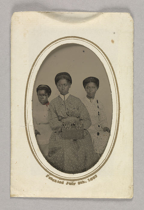 Tintype in a paper case of three unidentified young women seated, 1870s. Creator: Unknown. This tintype  a  depicts three  3  unidentified young African American women seated and facing the camera. The woman at center is slightly in front of the other two. She wears a dark colored dress with a lighter printed design and a pointed white collar. She holds a box or a thick book in her lap, and gilt is added to one of her left fingers and at the center of her collar. The woman at the right side wears a light colored overdress with dark floral print design and a light colored skirt with a ruffled hem. Gilt is added to an oval brooch she wears at her neck over a small rounded collar. The woman at the left side wears a white muslin blouse and apron over a light colored skirt. Gilt is added to one of her right fingers and a red bow is colored in at her neck. All three women wear their hair pulled up at the back. Their faces have been hand tinted. The reverse is blank. The tintype is placed inside a paper envelope  b  with an oval shaped window cut into one side. Gilt print lines the window and text below it reads  quot Patented July 8th, 1883 quot .