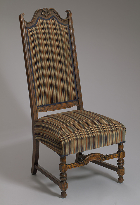 Upholstered side chair from Mae s Millinery Shop, 1900 1950. Creator: Unknown. High backed wooden side chair with an upholstered seat and back. The chair has early Baroque ball turned front legs and straight back legs. There is a carved scallop with feathered edges at the center of the front stretcher, the side stretchers are straight and there is no rear stretcher. The crest of the seat back has a carved late Baroque style shell design at the center and two inward facing ears on each side of the crest rail. The upholstery on the seat covers the front, side, and rear seat rails. The fabric is velvet with a striped design in red, yellow, orange, green, brown, beige, and black. The back upholstery is edged on the facing and reverse sides with blue passemenetrie with an undulating pattern. Mae Reeves  1912 2016  was a pioneering African American milliner who was famous for her custom made hats. Her business, Mae s Millinery Shop in downtown Philadelphia, was one of the first shops in the city to be owned by a woman of African American heritage.