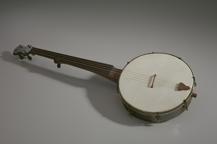 Banjo made in the style of William Esperance Boucher, Jr., ca. 1850s. Creator: Unknown. Banjo made in the style of William Esperance Boucher, Jr.  circa 1850s . The banjo has a circular head, with a tension head pulled tight across the front. There is a slightly tilted bridge on the head, with a tailpiece at the bottom holding the strings bottom in place along the rim. Both the tailpiece and the bridge are made of the same medium brown wood. There are metal brackets surrounding the rim of the banjo. This banjo is an early style of five string banjos, which has four full length strings alongside a short fifth string. The neck and fingerboard of the banjo is made of the same medium glossy wood as the back of the head. There are four wooden turning keys and pegs on the top, with a 5th string turning peg, also made of wood, about halfway up the neck or fingerboard.