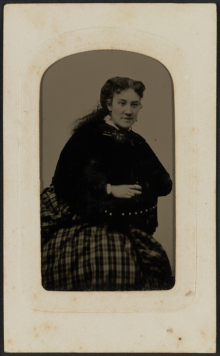Tintype portrait of Mrs. Annie Cox, ca. 1865. Creator: Hall s Tintype  amp  Ferrotype Gallery. Tintype of Mrs. Annie Cox shown in three quarter portrait. Cox is seated and leaning with her arms crossed on the arm of a chair. She is turned slightly to her left and looks off frame. Her curly hair is parted at the center and loosely gathered into a ponytail at the nape of her neck. She wears a dark bodice and a plaid skirt. There is a ring on her right hand ring finger. The tintype is housed in its original  quot Potters Patent quot  die cut paper frame. Information for Hall s Tintype  amp  Ferrotype Gallery is printed on the back of the photograph. The photograph is housed in the album 2017.30. The album page has a triple lined, gold border framing the print. Handwritten in graphite inside the bottom border of the printed frame of the album page is the text  quot Mrs. Anna Cox quot .