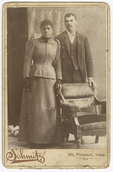 Photograph of an African American couple standing behind a chair, ca. 1890. Creator: Schmitz Studio. A black and white cabinet card photograph of a woman and man in formal dress standing next to a chair. The woman is wearing a dark colored skirt, top and dark fingerless gloves. Her left hand is resting on the chair back. The man is wearing a dark colored suit with a light colored shirt and bowtie. His left hand is resting on the chair back. Text printed on the card below the photograph says   x201c Schmitz  x201d  and   x201c Mt. Pleasant, Iowa.  x201d  There are no inscriptions.