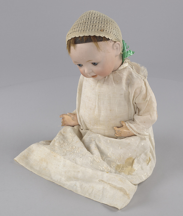 Doll owned by Clementine Roundtree Cottee and Josephine English Church, ca. 1920. Creator: Unknown. Doll representing a white baby, owned by African American children. Dolls representing black children were manufactured at this period, but were often  mammy doll  stereotypes. It is wearing a white cotton dress, two knit socks, a cotton undergarment and a knit cap. The dress is fastened with a safety pin and the cap ties with a green ribbon. The doll has blue glass eyes, rosy lips and cheeks, and short brown hair coming out from underneath the bonnet.