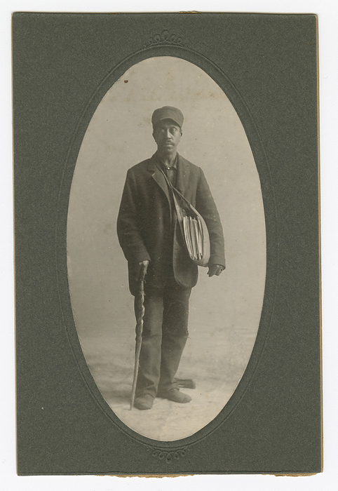 Cabinet card of a newspaper carrier, mid to late 19th century. Creator: Unknown. African American man standing in the center of the image against a light background. He is dressed in a dark hat and jacket, with a canvas newspaper carrier bag full of papers worn over his shoulder. In his proper right hand, he leans on a carved wooden walking stick. The photograph is cut to an oval shape and mounted on dark gray cardboard.
