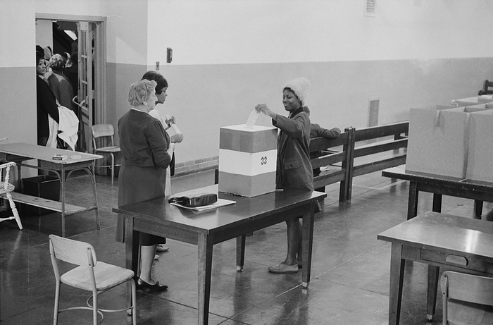 Young African American woman casting her vote in a ballot box at voting station in the Cardoza High School building, Washington. DC, 3 May 1964. Photographer:  Marion S Trikosko.