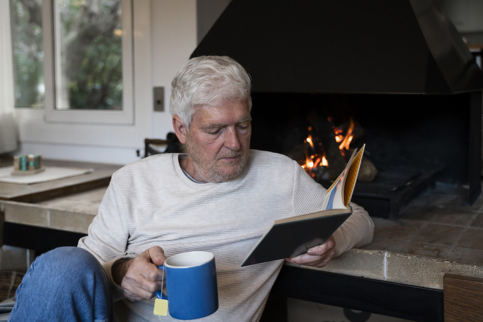 Barcelona, Spain. Mature man at home by the fireplace. Cozy, stay at home, relax, free time, active senior, leisure, hygge, relaxation, tranquility, retiree, pensionist, baby boomer Senior man with coffee cup reading book while sitting by fireplace at home