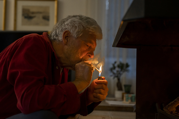 Barcelona, Spain. Mature man at home by the fireplace. Cozy, stay at home, relax, free time, active senior, leisure, hygge, relaxation, tranquility, retiree, pensionist, baby boomer Senior man igniting cigarette with lighter while sitting at home