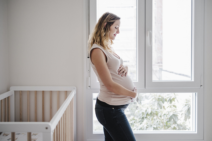 Pregnant woman with hands on stomach contemplating while standing by window at home