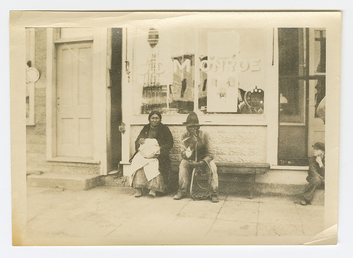 Photograph of a man and woman sitting outside of a storefront, early 20th century. Creator: Unknown. This gelatin silver print depicts a black and white image of a man and woman sitting on a bench in front of a store. The man is sitting on the right and wears a large hat, long sleeve shirt and pants. He is holding a harness in his hand. The woman next to him is holding a bundle of fabric in her lap. She is wearing a dark colored coat and a dress or skirt. They are both looking at the camera. The words painted on the store window behind them read  OLD MONROE . There are two hand drawn vertical lines on the back of the photograph.