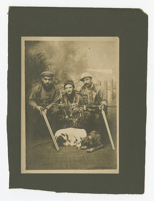 Photograph of a hunting party, ca. 1903. Creator: Unknown. A black and white photograph of a hunting party. There are three seated men holding game and rifles with two dogs lying at the feet of the men in the foreground. The photograph was taken in front of an outdoor backdrop with trees depicted on the left and a picket fence on the right. On the left side of the image is Will Westmoreland. He is wearing a newsboy cap, striped pants, a medium toned hunting jacket and a button down hunting vest with shell pockets on the front, holding ammunition. In the center is Thomas W. Holmes, Esq., wearing a dark shirt, rimmed cap and a medium toned vest and jacket. On the right side of the photograph is Will George in on the right wearing a light turtleneck shirt, white roughrider style hat and a medium toned hunting jacket and a button down hunting vest with shell pockets on the front, holding ammunition. The photograph has been mounted on dark grey cardboard. Several handwritten notes in black ink have been taped to the back that identifies the subjects.