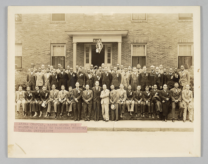 Photograph of Kappa Alpha Psi members, 1937 1938. Creator:  Dexheimer Carlon Studios. Members of the Kappa Alpha Psi fraternity in front of the Alpha Chapter house during the North Central Province regional meeting.  Kappa Alpha Psi founder and 1st Grand Polemarch Elder Watson Diggs is in the front row center, with his arms crossed. Directly behind him is John L. Stewart. Other prominent Kappa officials pictured standing beside Elder Watson Diggs from left to right are: 4th Grand Polemarch W. Ellis Stewart, 2nd Grand Polemarch Irven Armstrong, 9th Grand Polemarch Theophilus Mann, and to Diggs  x2019  right North Central Province Polemarch Julius Morgan standing and Grand Strategus Albert Spurlock seated. A typed label on pink paper that was matted with the photograph reads  ALPHA CHAPTER, KAPPA ALPHA PSI   FRATERNITY HOST TO REGIONAL MEETING   INDIANA UNIVERSITY .