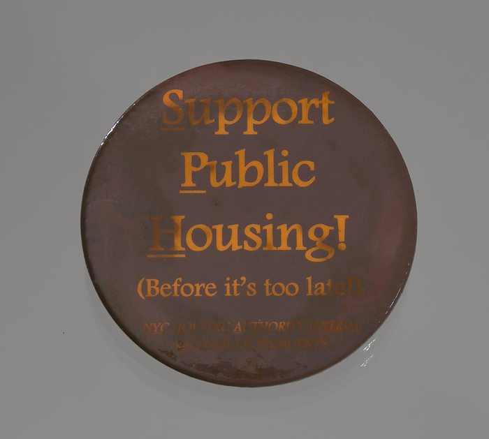 Pinback button promoting public housing in New York City, late 20th century. Creator: Unknown. An aubergine colored pinback button promoting public housing. The text appears in orange and reads:  Support   Public   Housing   with the first letter of each word underlined. Below this reads:   Before it s too late     NYC HOUSING AUTHORITY INTERIM   COUNCIL OF PRESIDENTS . The button is heavily stained and discolored. The back of the button has some scratches and stains and has a metal fastener and clasp.