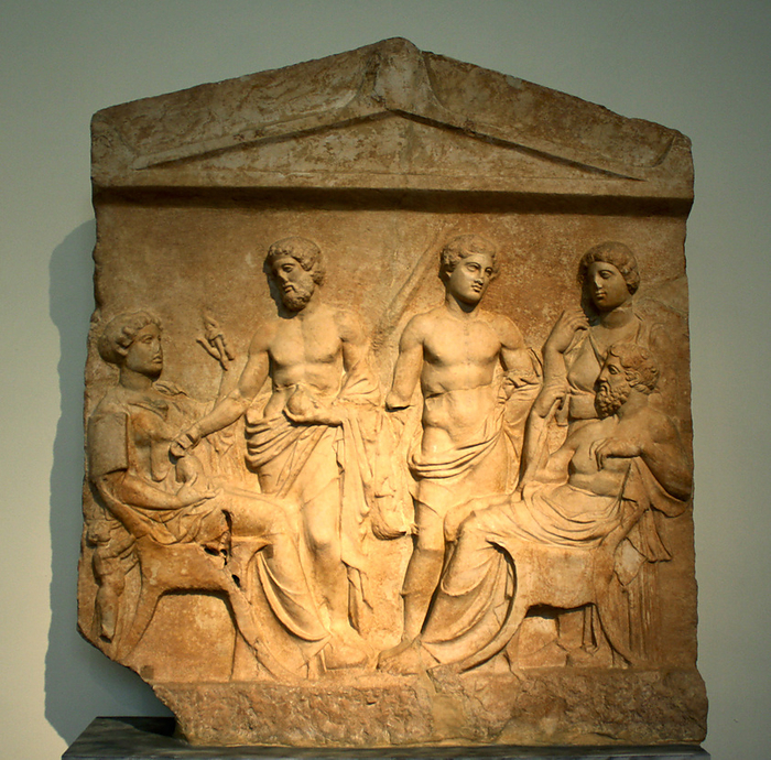 Grave stele.  Thespian marble.  Found in the outskirts of Thebes.  Two figures, a bearded man with a staff on the right and a spinning (?) woman on the left, sit facing each other.  Three more figures two men and a woman stand in the background. A very small female attendant is represented at far left. Characteristic attributes are the pomegranate and torch (?) held by the standing bearded man as well as the aryballos in the right hand of the beardless youth.  End of the fifth century BC
