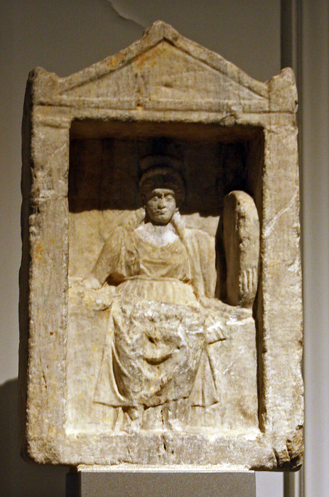 Marble votive relief with Cybele enthroned.  Post-2nd century AD.  The Goddess is represented in a small temple, wearing a sleeveless, girdled chiton, a himation and a high polos.  She holds a phiale in the right hand and a drum in the left.  Visible on her knees are traces of a lion.  The lion and the drum, which was played during rites in Cybele's honour, were the attributes of the Great Mother Goddess of Asia Minor.  Small temples of this kind, which perhaps denote the shallow caves in which statues of her were placed, are identified in sanctuaries of Cybele and other deities after the Late Classical period, in private houses and in cemeteries.
