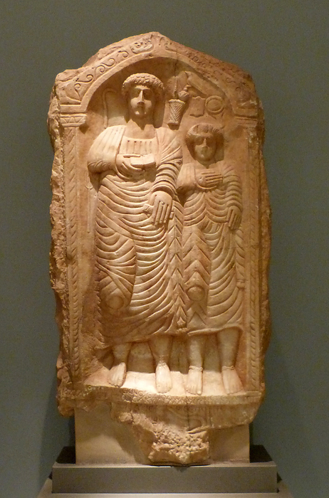 Marble inscribed grave relief. Represented is a couple in an arched conch, framed by the symbols of male diligence and female orderliness. The inscriptions attest the resilience of the Greek language.  From, Phrygia, Asia Minor, early 3rd century, AD