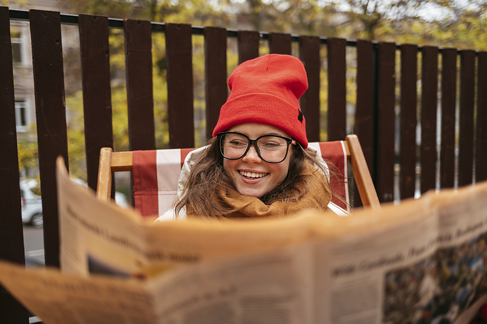 female Smiling woman wearing knit hat and eyeglasses reading newspaper while sitting at sidewalk cafe