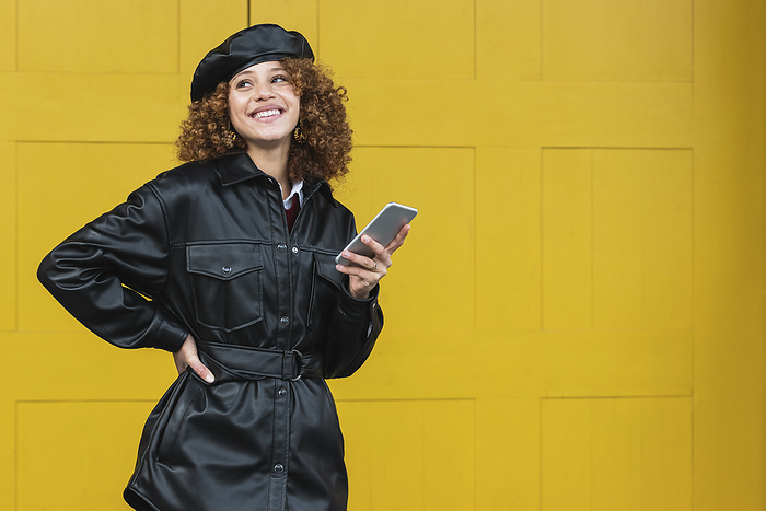 female Smiling woman wearing leather jacket and beret standing with mobile phone against yellow door