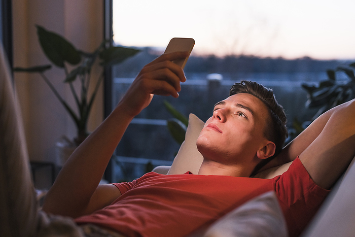 Young man looking at smartphone Relaxed young man lying on sofa while using smart phone in living room