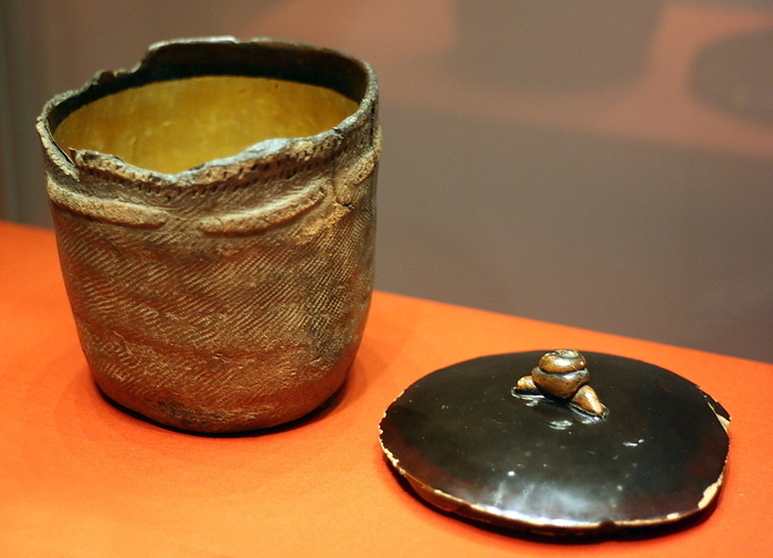 Jomon pot. This jomon cooking pot was about 2000 years earlier then flame and crown pots in the centre of the room. The rim is decorated with marks incised with a stick or finger nail and the cord markings are clearly visible. It was probably uncovered by a farmer in the 19th century and spotted by a tea master who thought it would make a fine water vessel (mizusashi) for a tea gathering. Gold leaf and lacquer was applied to the interior of the prehistoric vessel and a wodden lid was constructed with a snail decoration, alluding to the pot's unearthing from the soil.