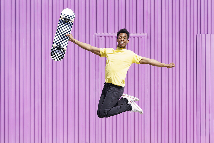 Cheerful man with arms outstretched holding skateboard whole jumping in purple corrugated iron