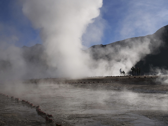 Steaming geyser, El Tatio, Chile A large plume of steam from one of the numerous geysers is illuminated by the early morning sunlight with a group of tourists taking photographs at El Tatio geothermal field high in the Andes of northern Chile. The surface is very fragile and stones are used to mark safe pathways through the field., Photo by DAVID TAYLOR SCIENCE PHOTO LIBRARY