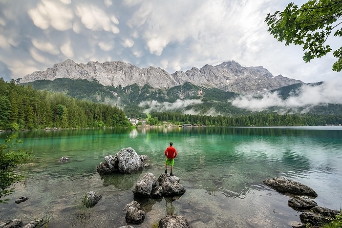 Young man standing on a rock on the shore, Eibsee lake in front of Zugspitze massif with Zugspitze, dramatic Mammaten clouds, Wetterstein range, near Grainau, Upper Bavaria, Bavaria, Germany, Europe