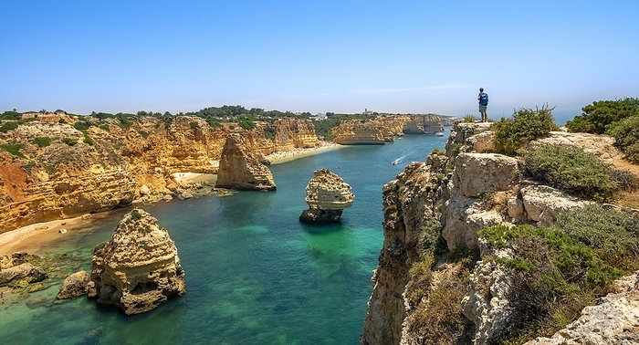 Young man standing on rocks on cliff, view on cliff of sandstone rocks, rock formations in turquoise sea, sandy beach, Praia da Marinha, Algarve, Lagos, Portugal, Europe