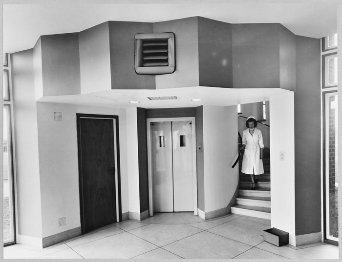 Shell Haven Oil Refinery, Shell Haven, Stanford Le Hope, 13 05 1957. Creator: John Laing plc. The ground floor entrance to the new administration building at Shell Haven Refinery, showing the circular lift tower and a woman walking down the winding tower staircase. The caption beneath this album print reads  Shell Haven Administration Block. Ground floor entrance, in the background is the lift tower which is circular and houses the electric passenger lift. On the right, the ground floor entrance to the circular staircase. The top section of picture shows the plaster feature in which the air conditioning louvre can be seen, and this octagonal feature gives an idea what the outside of the tower looks like the brickwork being similar. 
