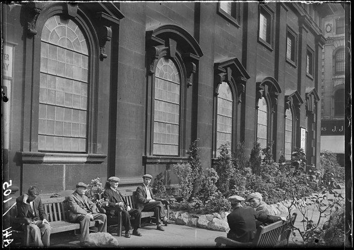 Holy Trinity Church, Boar Lane, Leeds, 1941. Creator: George Bernard Wood. Holy Trinity Church, Boar Lane, Leeds, 1941. An exterior view of the Holy Trinity Church, showing the south front with men sat on benches in the foreground . The church was built between 1721 and 1727 by William Etty. The upper tower was added after 1839 by R. D. Chantrell. It has a rectangular plan with seven by three bays, with a tower in the middle bay of the west end. The south front has a door in the first bay, windows with alternating triangular and segmental pediments on the ground floor, and a second tier above.