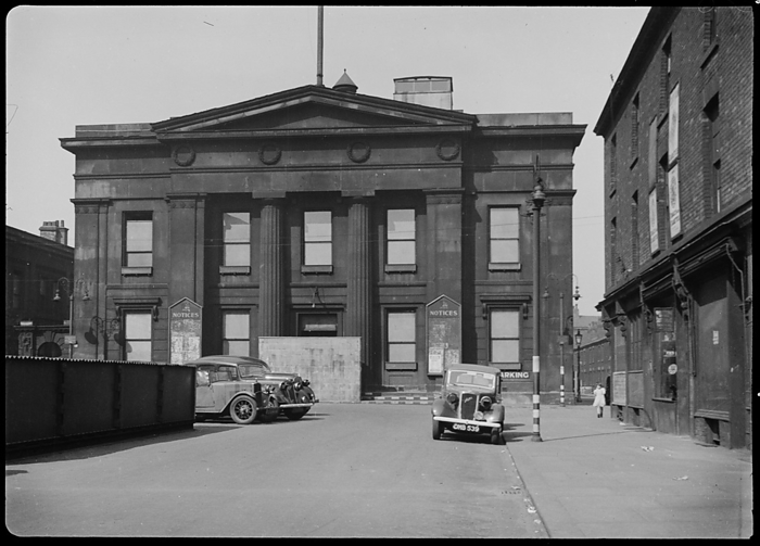 Town Hall, Bexley Square, Salford, 1942. Creator: George Bernard Wood. Town Hall, Bexley Square, Salford, 1942. An exterior view of the Town Hall, showing the south facade with two contemporary cars in the foreground. The hall was built by Richard Lane between 1825 and 1827, and the south facade has two storeys and five bays, with a three bay Doric portico with giant columns, a pediment and frieze with four relief wreaths. In the image the windows have blackout fabric covering them, and a temporary brick wall over the main entrance. The street furniture is painted with black and white stripes, to faciliate night time driving during World War II.