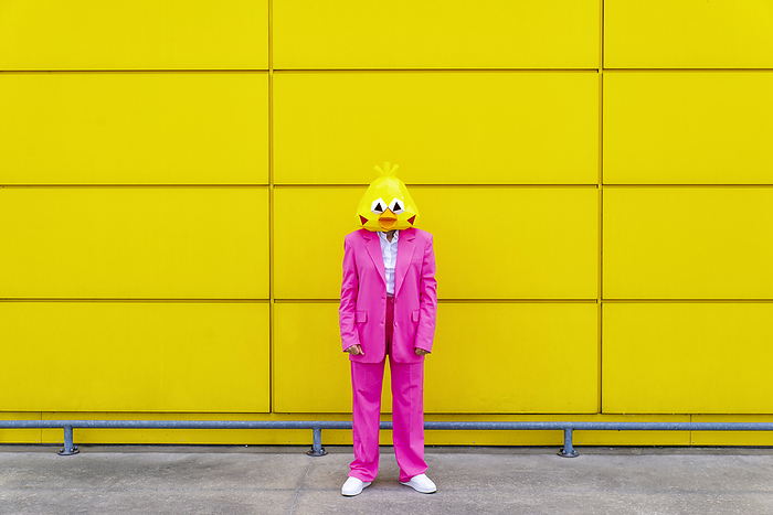 female Woman wearing vibrant pink suit and bird mask standing in front of yellow wall