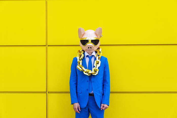 Man wearing vibrant blue suit, pig mask and large golden chain standing in front of yellow wall