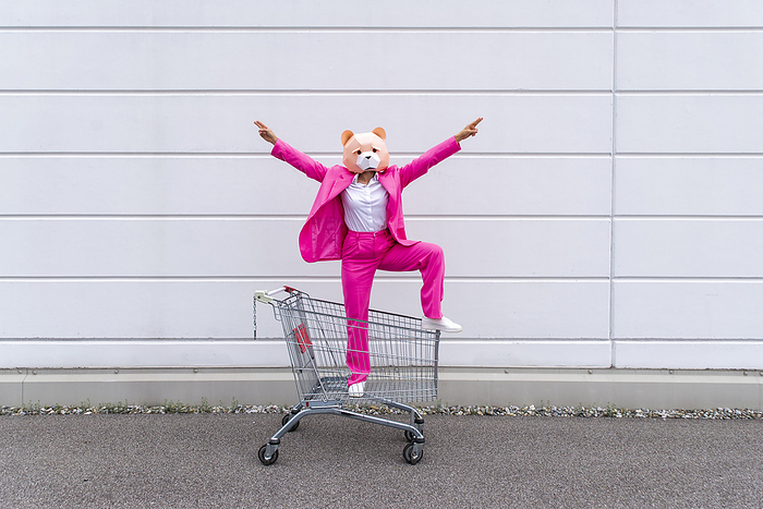female Woman wearing vibrant pink suit and bear mask standing in shopping cart with raised arms