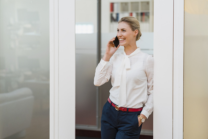 Cheerful female professional talking on smart phone while leaning at doorway in office