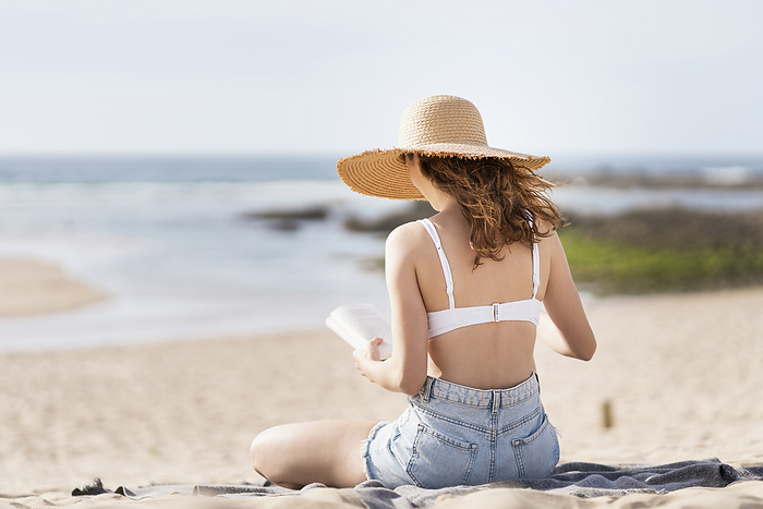 female Woman wearing sun hat reading book while sitting at beach during vacations