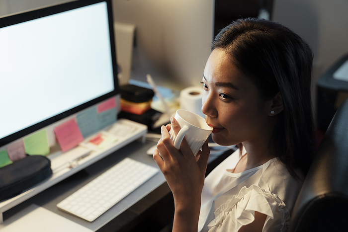 businesswoman Asian thai woman drinking and sipping hot coffee from a cup, sitting infront of computer at office. Night shift time.